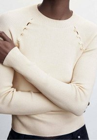 Rippato cremefarbener Pullover mit rundem Ausschnitt und dekorativen Knopfakzenten an den Schulternähten. Lange Ärmel mit eng anliegenden Bündchen.