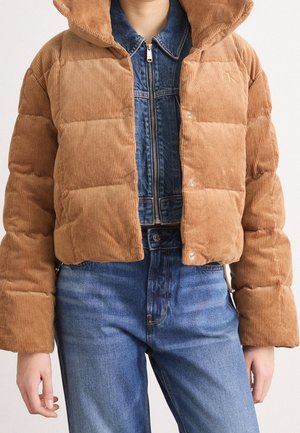 Personne portant une veste courte matelassée en velours côtelé beige par-dessus une veste en jean bleue et un jean bleu, debout, les bras relâchés.