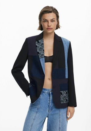 Patchwork blazer with black, blue denim, and textured fabric. Two buttons, lapel collar, and front pockets. Contrast paneling details.
