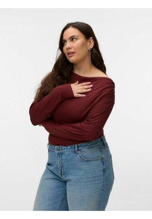 Ženska z dolgimi rjavimi lasmi, oblečena v bordo majico in modre kavbojke, z eno roko na prsih, gleda vstran.