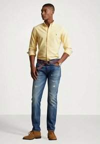 Mlad moški na manjšem vasovanju predstavlja sproščeno oblačilo z rumeno zapnjenim srajcem, strganimi modrimi kavbojkami, črtastim pasom, rjavimi semiš čevlji in zapestnico.