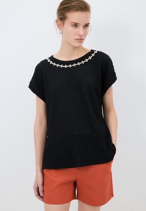 Haut noir texturé à manches courtes, avec un col décoratif en perles blanches. Associé à un short orange.