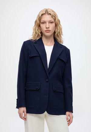 Une femme blonde porte un blazer bleu marine à un bouton sur un haut blanc et un pantalon crème, sur un fond clair et uni.