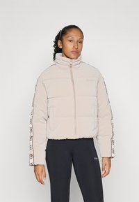 Kvinne står iført en lysebeige dunjakke med logobånd på ermene og svarte leggings mot en ensfarget hvit bakgrunn.
