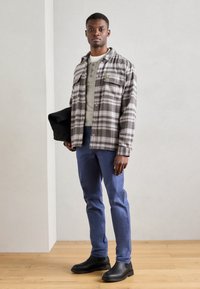 Grau-weißer karierten Flanellhemd, darunter ein hellgraues Henley, blaue Chinos, schwarze Stiefeletten, hält eine schwarze Tasche. Holzboden im Hintergrund.