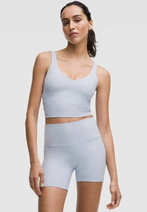 Mladá žena v světle šedé sportovní podprsence a vysokých cyklistických šortkách, stojící na jednobarevném šedém pozadí.