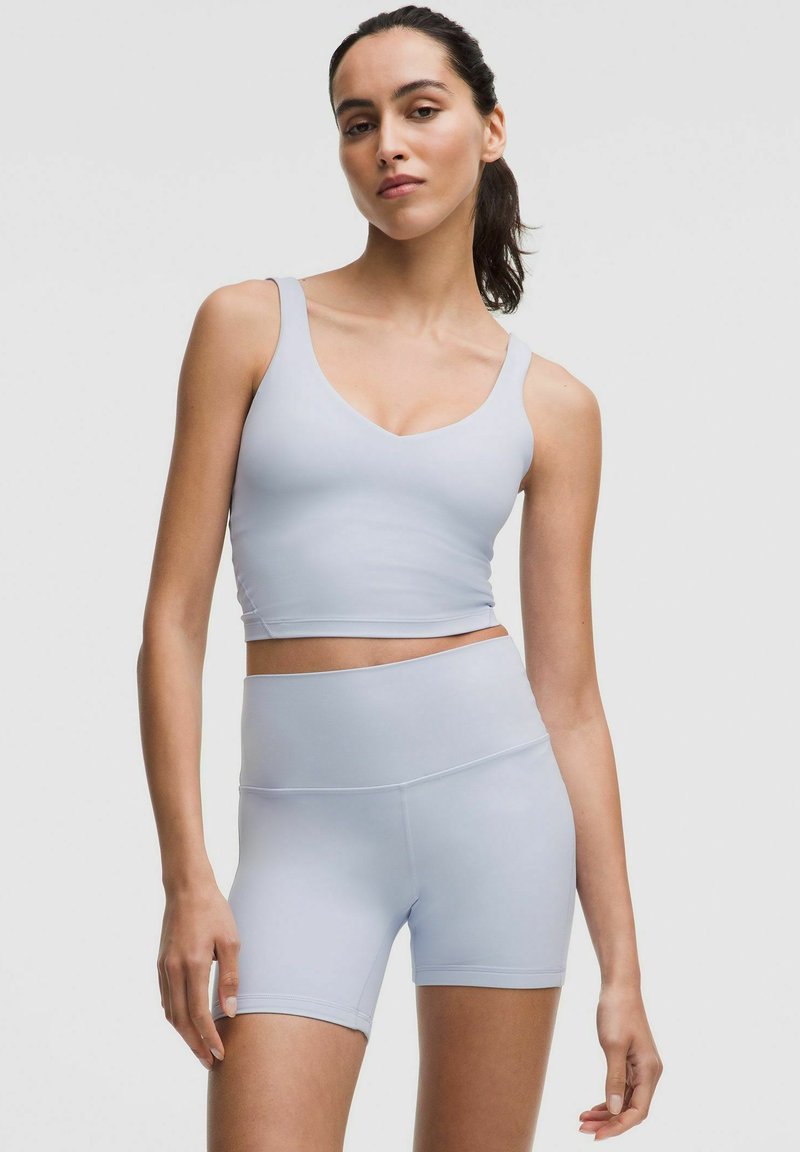 Giovane donna che indossa un reggiseno sportivo grigio chiaro e pantaloni da ciclista a vita alta, in piedi contro uno sfondo grigio uniforme.