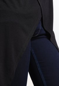 Camicetta nera con orlo curvo, realizzata in materiale morbido, indossata sopra un paio di jeans blu scuro con cuciture a contrasto lungo le cuciture.