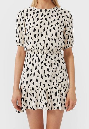 Vrouw draagt een korte, crèmekleurige jurk met zwarte onregelmatige vlekken, met pofmouwen, elastische taille en gerimpelde zoom.