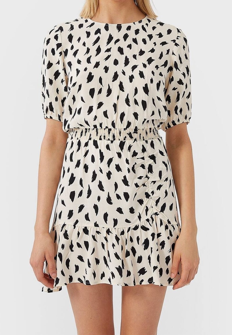 Femme portant une robe courte crème avec des taches noires irrégulières, à manches bouffantes, taille élastique et ourlet volanté.