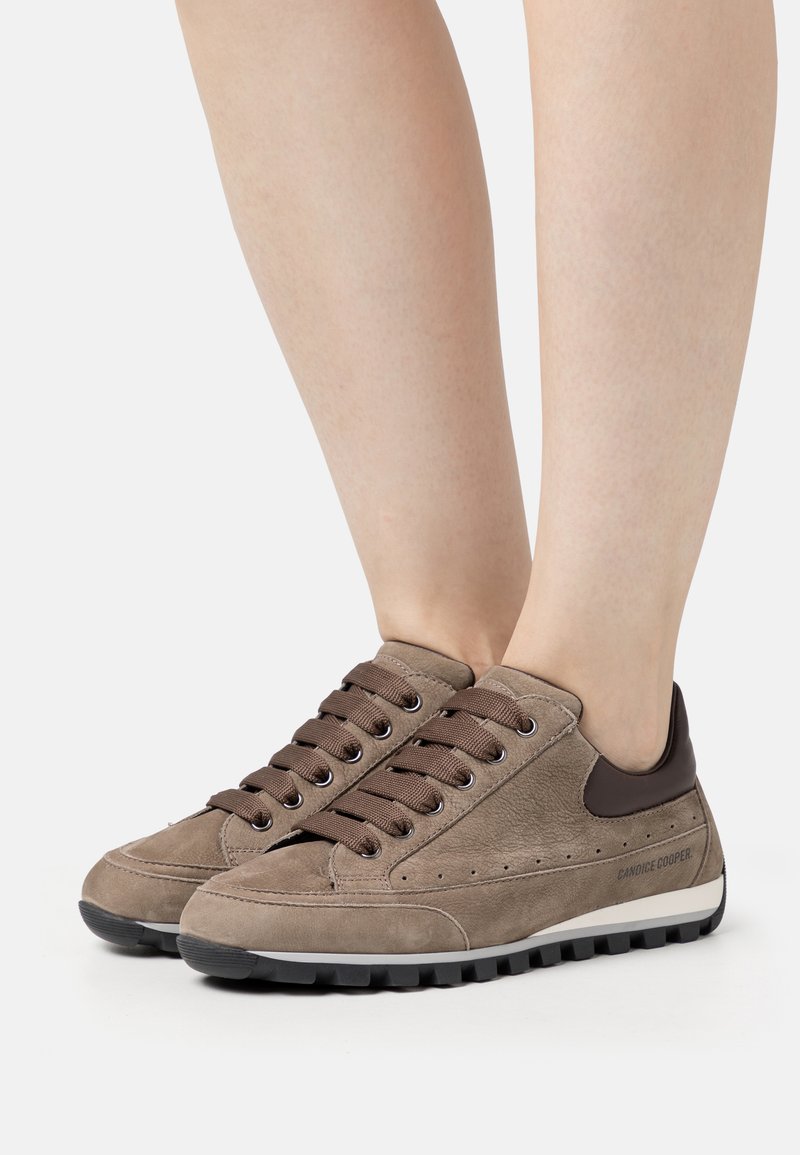 Des baskets en daim marron présentent un bout arrondi, des lacets marron et une semelle en caoutchouc noir contrastante. Des accents latéraux affichent de petites perforations.