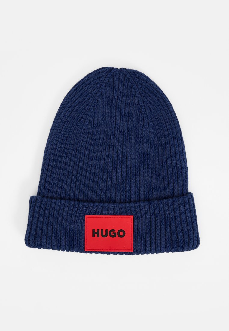 Marineblauer gestrickter Beanie mit gerippter Textur, ausgestattet mit einem umgeschlagenen Bündchen und einem roten rechteckigen Patch mit dem Wort "HUGO" in Schwarz.