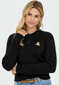 Juodas megztas polo megztinis su pynių raštu, ilgomis rankovėmis, trijų mygtukų užsegimu ir maža siuvinėta meškiuko detalė krūtinėje.