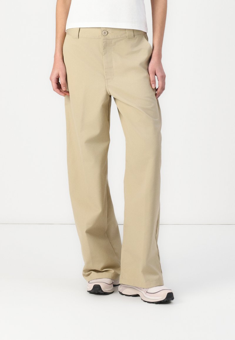 Pantalons larges beige en matériau léger avec une fermeture à bouton et deux poches avant, présentant une texture lisse et des lignes épurées.