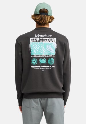 Personne portant un sweat-shirt noir avec des motifs et un texte aventure en bleu sarcelle et blanc au dos, associé à un pantalon gris et une casquette verte.