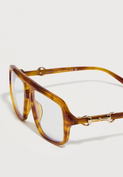 Τετράγωνα γυαλιά με θήκη από χελώνα και διαφανείς φακούς, με χρυσή λεπτομέρεια σε σχήμα αλόγου στις πλευρές και την επιγραφή «GUCCI Made in Japan» στο εσωτερικό του βραχίονα.