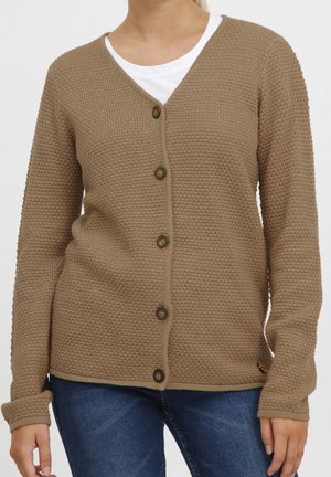 Femme portant un cardigan marron texturé boutonné sur une chemise blanche et un jean bleu, debout devant un fond uni.