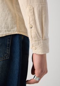 Beige shirt with textured fabric, buttoned cuffs, and a slightly curved hem. Dark denim pants visible below. Hand adorned with a silver ring.