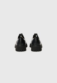 Chaussures en cuir noir avec une semelle épaisse, dotées d'un bout arrondi et d'un accent arrière avec logo. La semelle présente des rainures profondes et prononcées.