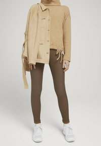 Chaqueta de ante beige sobre un suéter de punto color canela con cordón, combinada con leggings marrones y zapatillas blancas.