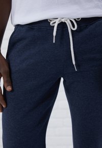 Marina joggers i mjukt tyg, med dragsko i midjan och vit snodd med silverfärgad spets. Närbild med en hand som vilar på benet.