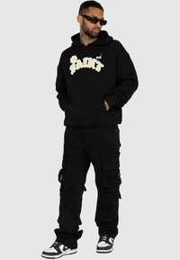 Hoodie noir avec le logo blanc "SAINT", associé à un pantalon cargo noir et des baskets noir et blanc. Des lunettes de soleil complètent le look.