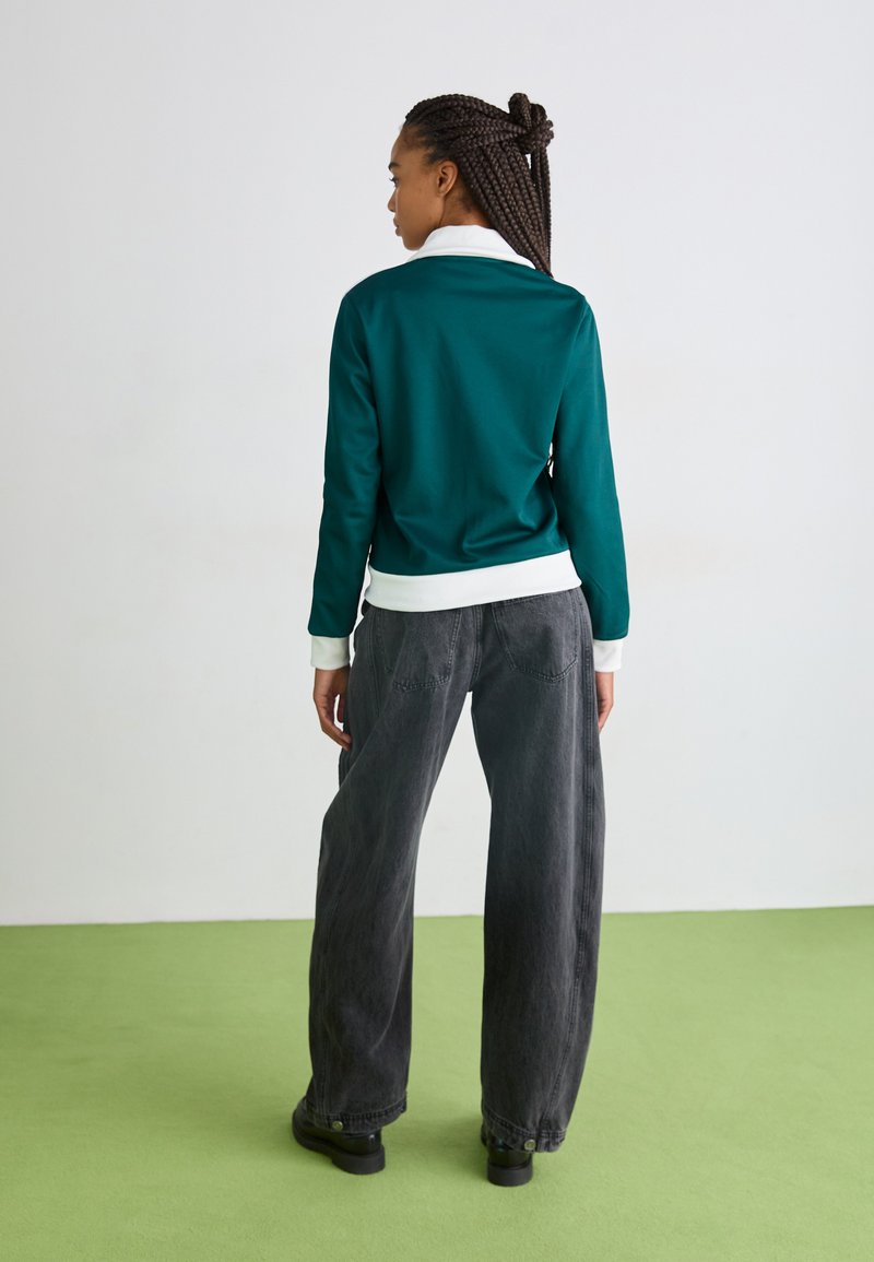 Türkiser Sweatshirt mit einem weißen Kragen, kombiniert mit weiten grauen Jeans, vor einem hellen Hintergrund und grünen Boden ausgestellt.
