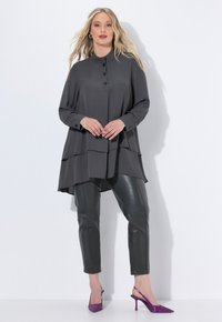 Woman wearing a layered charcoal tunic, black leather pants, and purple pointed-toe heels, standing against a white background.