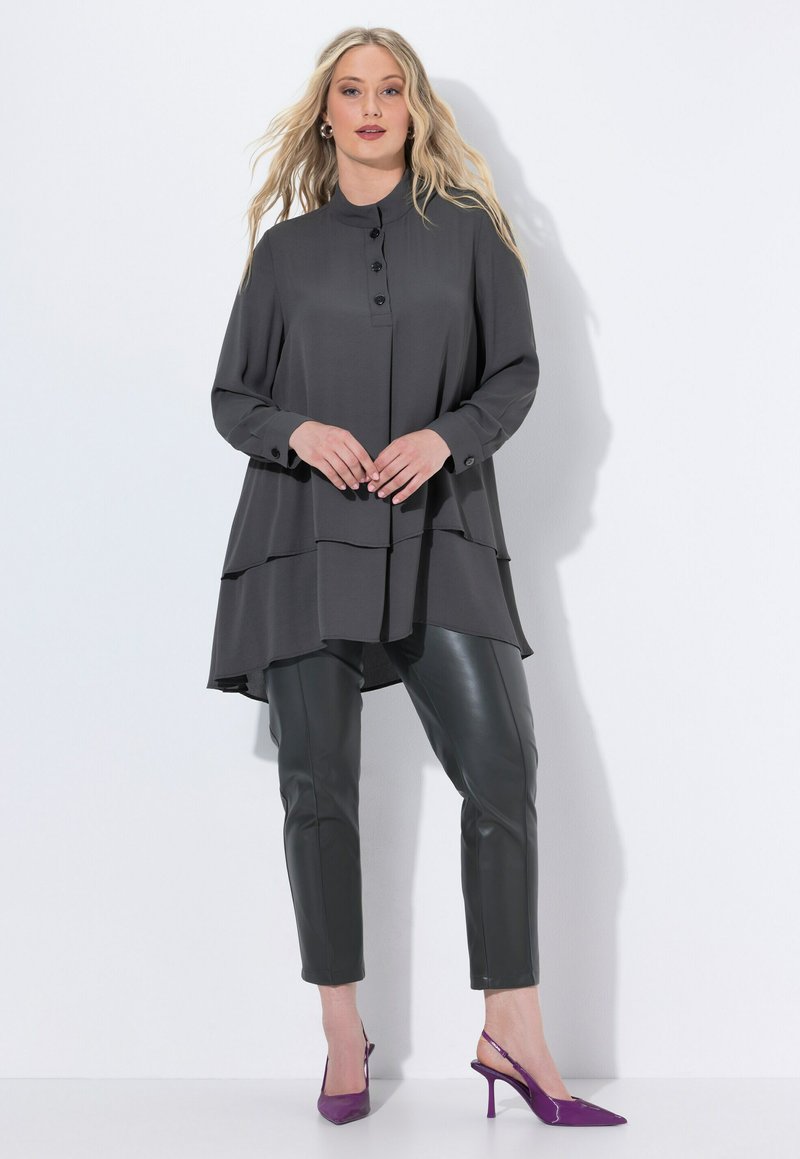 Woman wearing a layered charcoal tunic, black leather pants, and purple pointed-toe heels, standing against a white background.