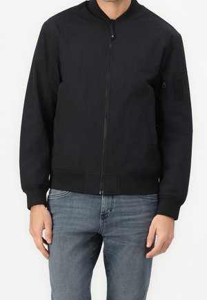 Mann trägt schwarze Bomberjacke mit Reißverschluss und blaue Jeans, steht mit entspannten Händen an den Seiten.
