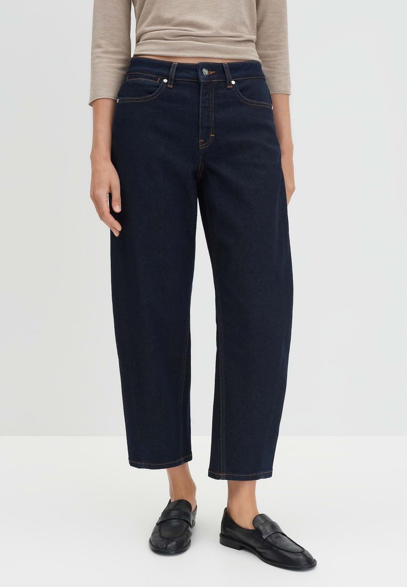 Jeans a gamba larga in denim un blu scuro con vita alta, caratterizzati da cuciture arancioni e tasche standard, abbinati a scarpe slip-on in pelle nera.