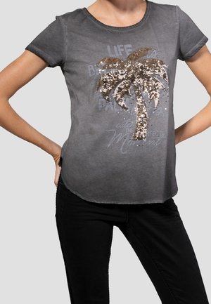 Frau trägt ein graues kurzärmeliges Shirt mit einem glitzernden Palmenmotiv und verblasstem Schriftzug, kombiniert mit schwarzer Hose, Hände in die Hüften gestützt.