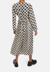 Dlouhé rukávy šaty v černobílém vzoru, s těsným pasem a páskem, splývavou sukní a texturovanou tkaninou. Černé boty dotvářejí vzhled.