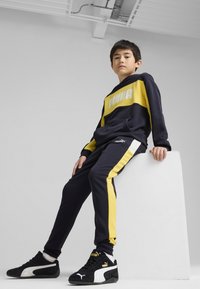 Granatowy i żółty hoodie Puma z logo, zestawiony z granatowymi spodniami dresowymi z żółtymi paskami po bokach. Czarne sneakersy z białymi akcentami.