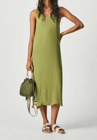 Žena v bezrukávových olivově zelených midi šatech drží prošívanou olivově zelenou kabelku s béžovým popruhem, obutá v béžových sandálech na přezku, stojící.