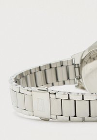 Tuva skatu uz sudraba metāla pulksteņa siksniņu ar salokāmu sprādzi un iegravētu taisnstūra logo uz baltas fona.