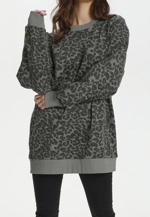 Frau trägt ein locker sitzendes, graues, langärmeliges Oberteil mit Leopardenmuster, gerippten Bündchen und Saum, kombiniert mit schwarzen Hosen, stehend vor weißem Hintergrund.