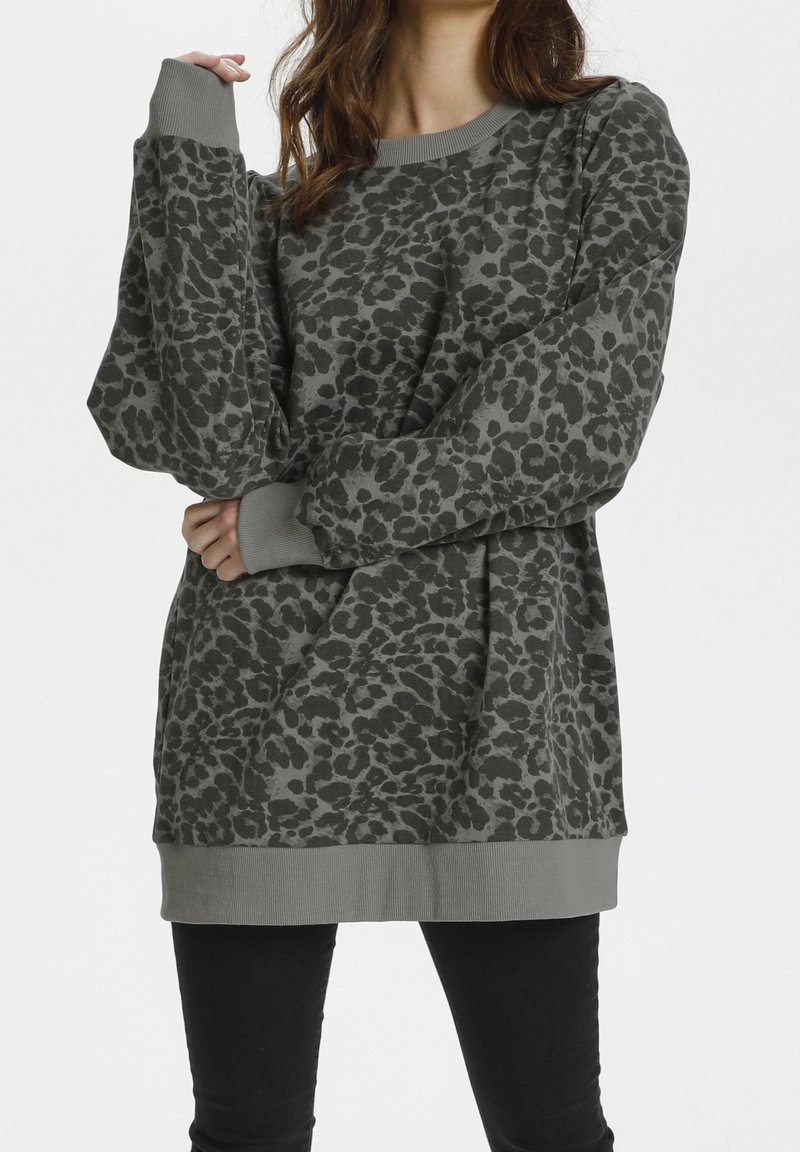 Femme portant une chemise ample à manches longues gris imprimé léopard avec poignets et ourlet côtelés, associée à un pantalon noir, debout devant un fond blanc.