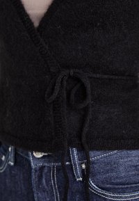 Svart strikket omslagstopp med V-hals, bundet i midjen med en myk tekstur. Båret over en lysfarget topp og kombinert med mørkeblå jeans.