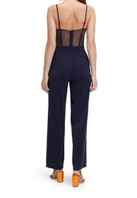 Femme portant une combinaison bleu marine avec une partie arrière transparente et des bretelles spaghetti, associée à des sandales à talons blocs métalliques orange.