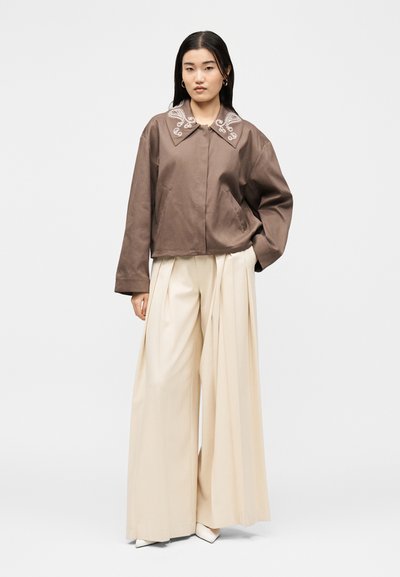 Νεαρή γυναίκα που φοράει φαρδιά κρεμ παντελόνια και ένα φαρδύ καφέ σακάκι με κεντημένο γιακά, με τα χέρια στις τσέπες, σε ανοιχτό φόντο.