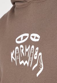 Hoodie en coton marron avec un design graphique blanc représentant des yeux et des mains, affichant le mot "KARMA" en évidence.