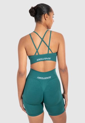 Teal sportbeha en high-waisted shortset met gekruiste banden, textuurstof en logo-detail op de tailleband.