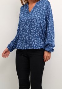 Blaue Bluse mit Blumenmuster, V-Ausschnitt, langen Ärmeln und gerafften Manschetten; leichtes Material mit kleinen, hellblauen Blumenmustern überall.