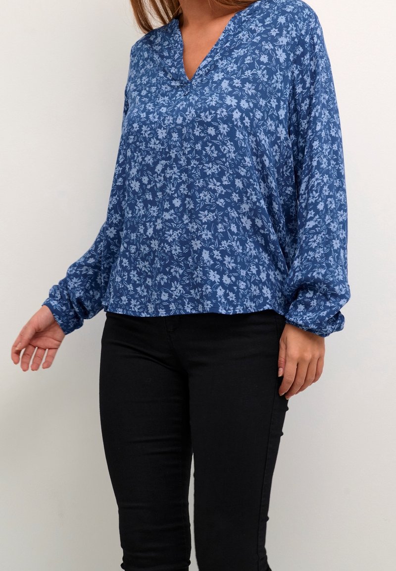 Blaue Bluse mit Blumenmuster, V-Ausschnitt, langen Ärmeln und gerafften Manschetten; leichtes Material mit kleinen, hellblauen Blumenmustern überall.