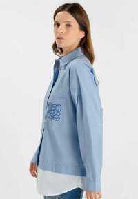 Lichtblauw gestreept shirt met een kraag, voorzien van een zak en blauwe geborduurde grafische details op de borst. Lange mouwen en een ontspannen pasvorm.