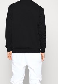 Sudadera negra de mezcla de algodón, con cuello redondo y puños acanalados. Combinada con pantalones blancos, mostrando un diseño trasero simple.