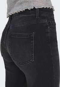 Gros plan d'une personne portant un jean en denim noir et un haut gris côtelé, axé sur la poche arrière et la zone de la taille.