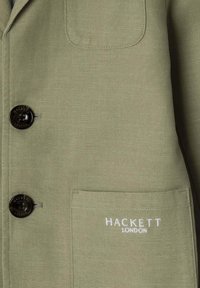 Oliwkowy żakiet z teksturowanej tkaniny, z dwoma czarnymi guzikami z wytłoczonym napisem "Hackett London" oraz lewą kieszonką na piersi z białym haftem.