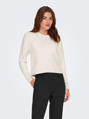 Femme portant un pull crème à manches longues avec de petites décorations et un pantalon noir, debout avec une main dans la poche sur fond blanc.